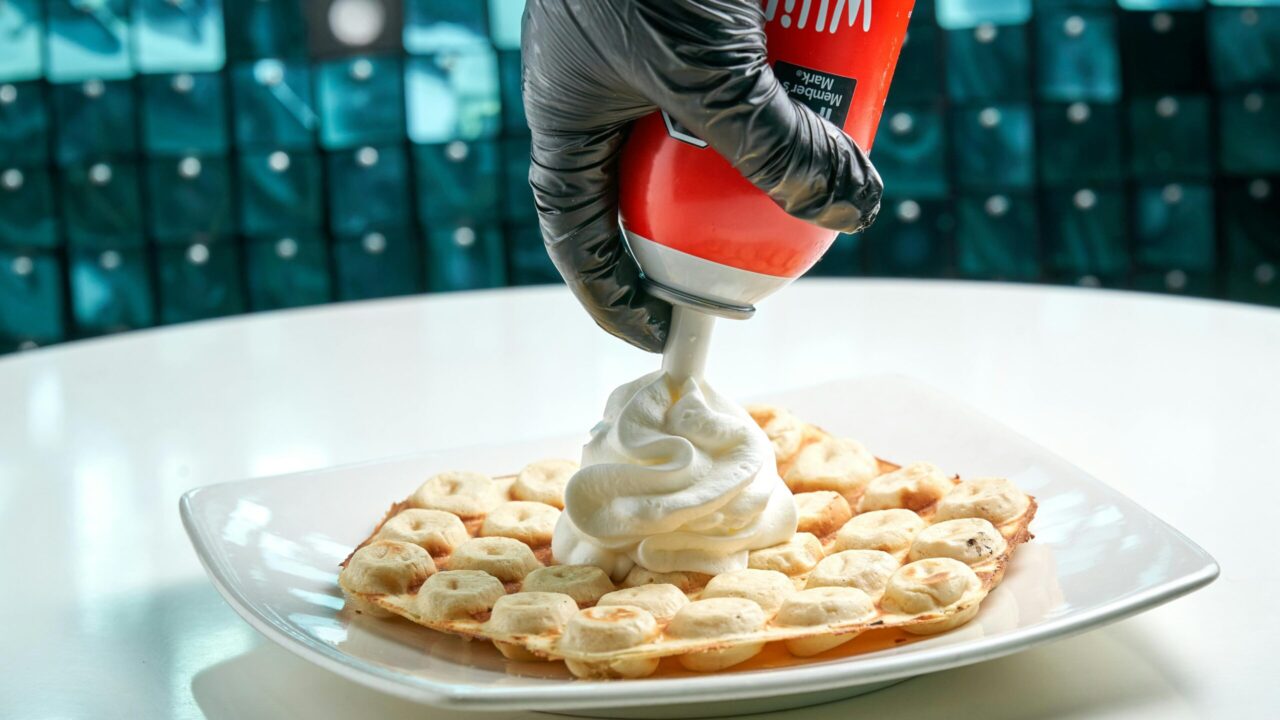 Close-up of a waffle being topped with whipped cream by a gloved hand.